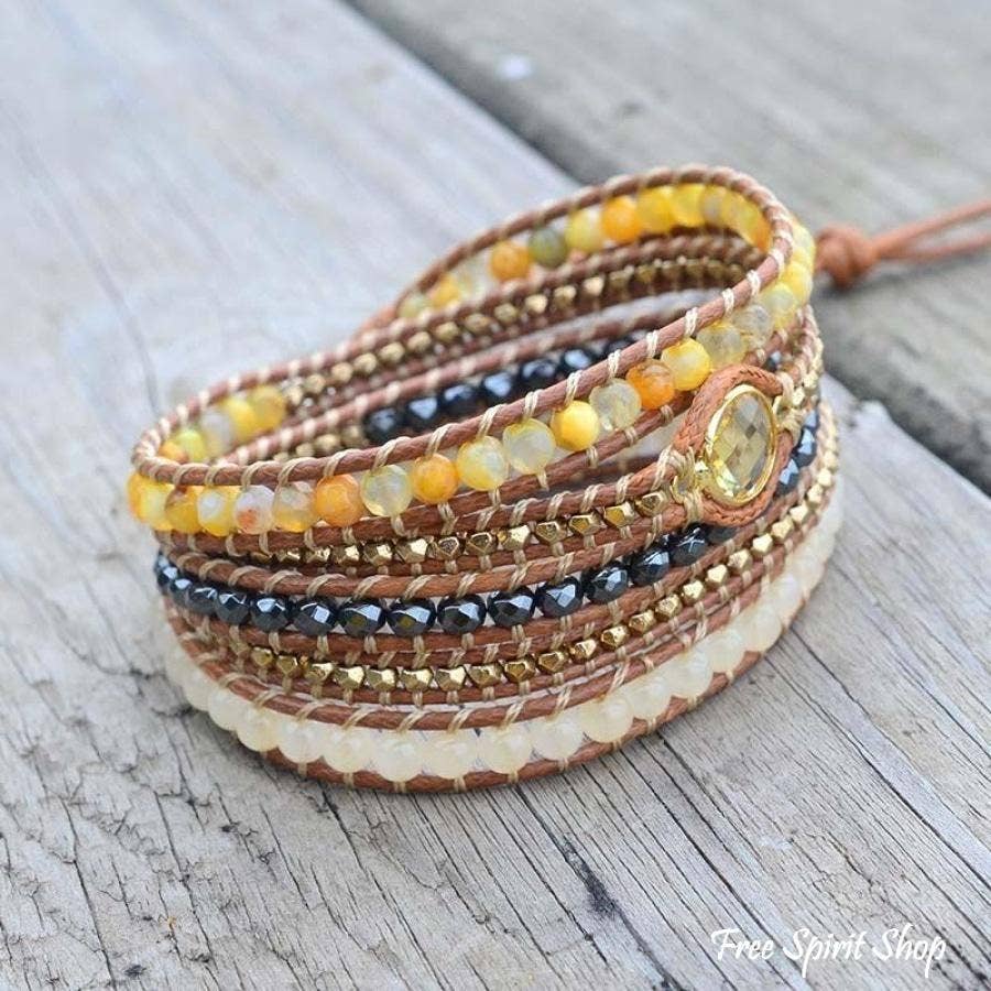A multi-layered wrap bracelet with brown leather cords, yellow and orange beads, black faceted beads, gold spacers, and a round gold-toned charm, displayed on a weathered gray wooden surface.