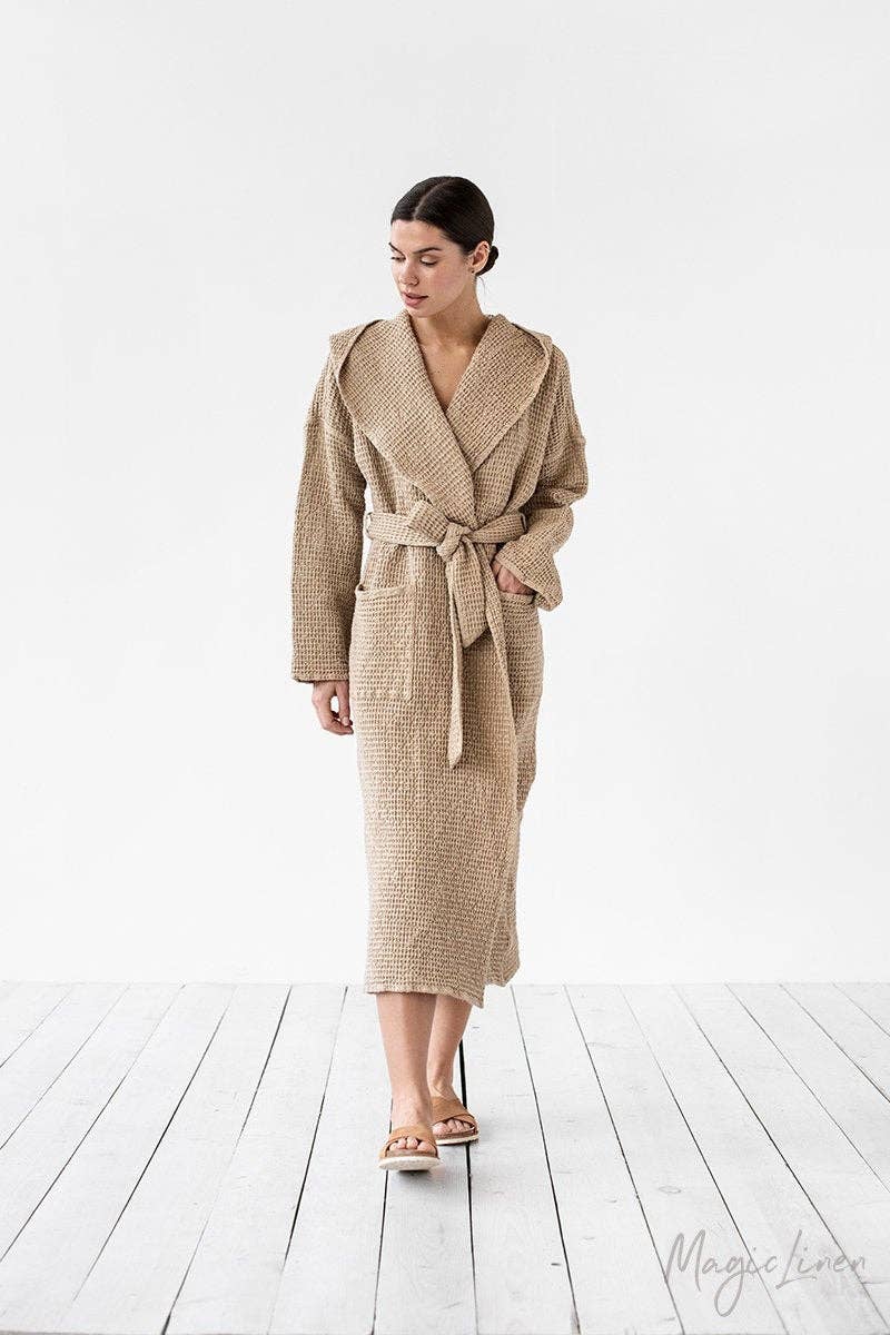A woman modeling a beige textured waffle-knit robe with a tie waist belt, standing on a light gray wooden plank floor against a white studio background.