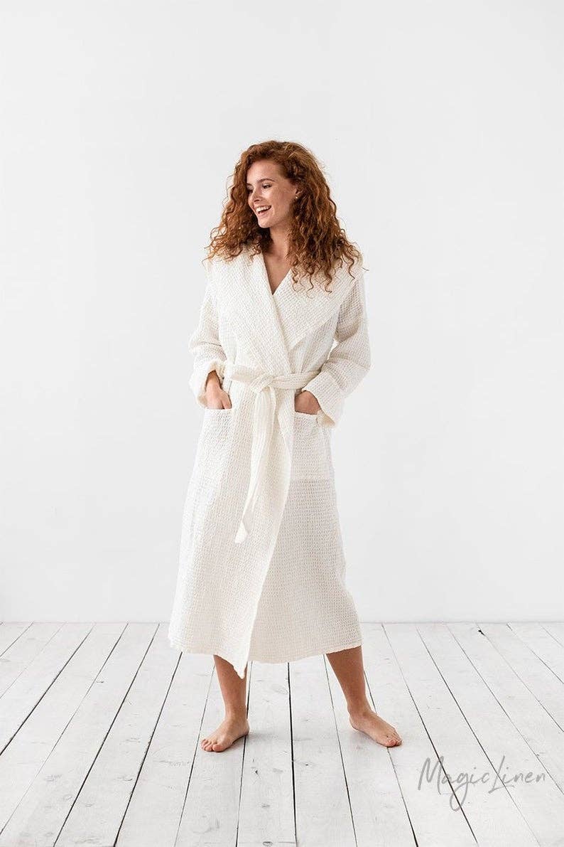 A woman with curly red hair wearing a cream-colored textured bathrobe, standing barefoot on a white wooden plank floor against a white studio background.