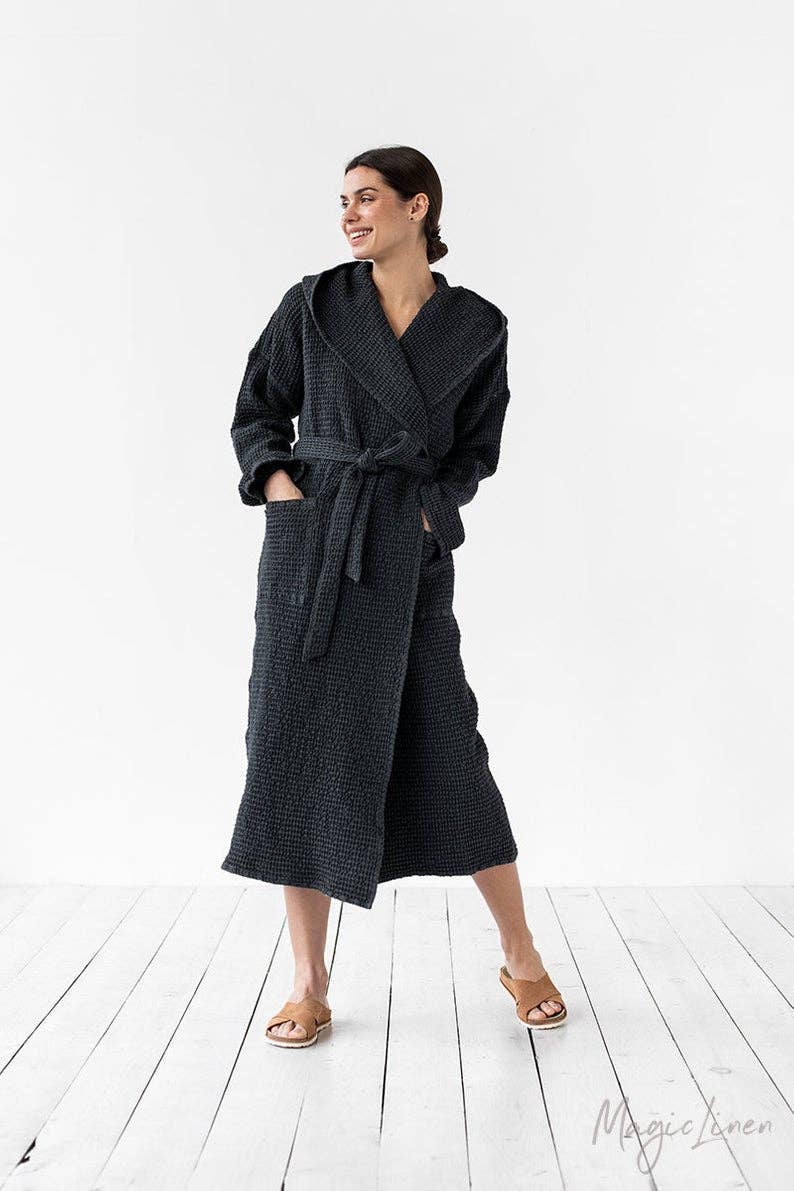 A woman modeling a dark charcoal gray textured waffle-weave bathrobe with a tie waist, standing on a white wooden plank floor against a white studio background.