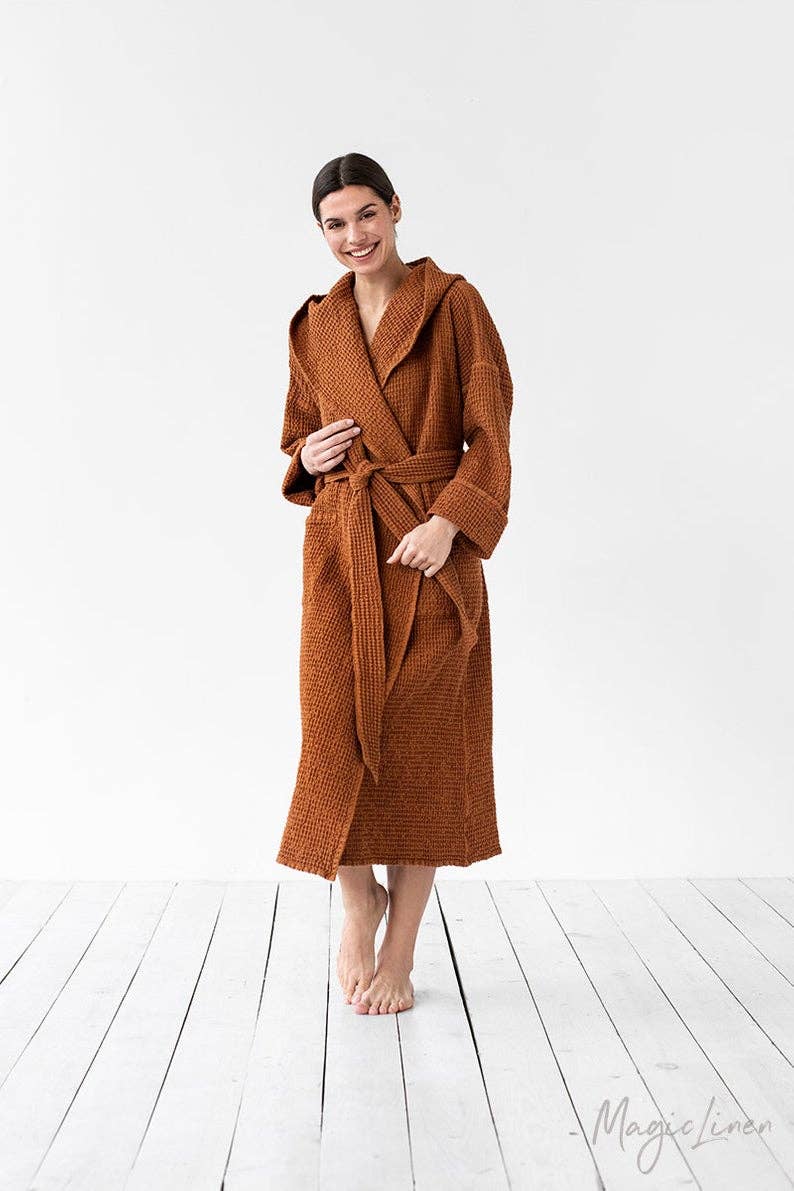 A woman stands barefoot in a studio wearing a waffle-knit bathrobe in a warm rust brown color. The robe has a shawl collar, long sleeves, and a matching waist tie.