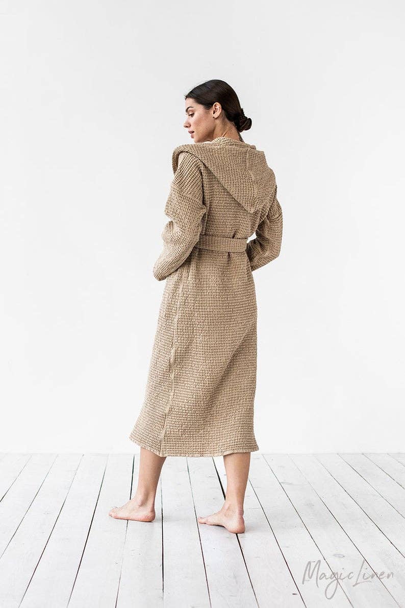 A woman stands barefoot in a studio wearing a hooded waffle-knit robe, shown from the back. The robe is a warm taupe/tan color.