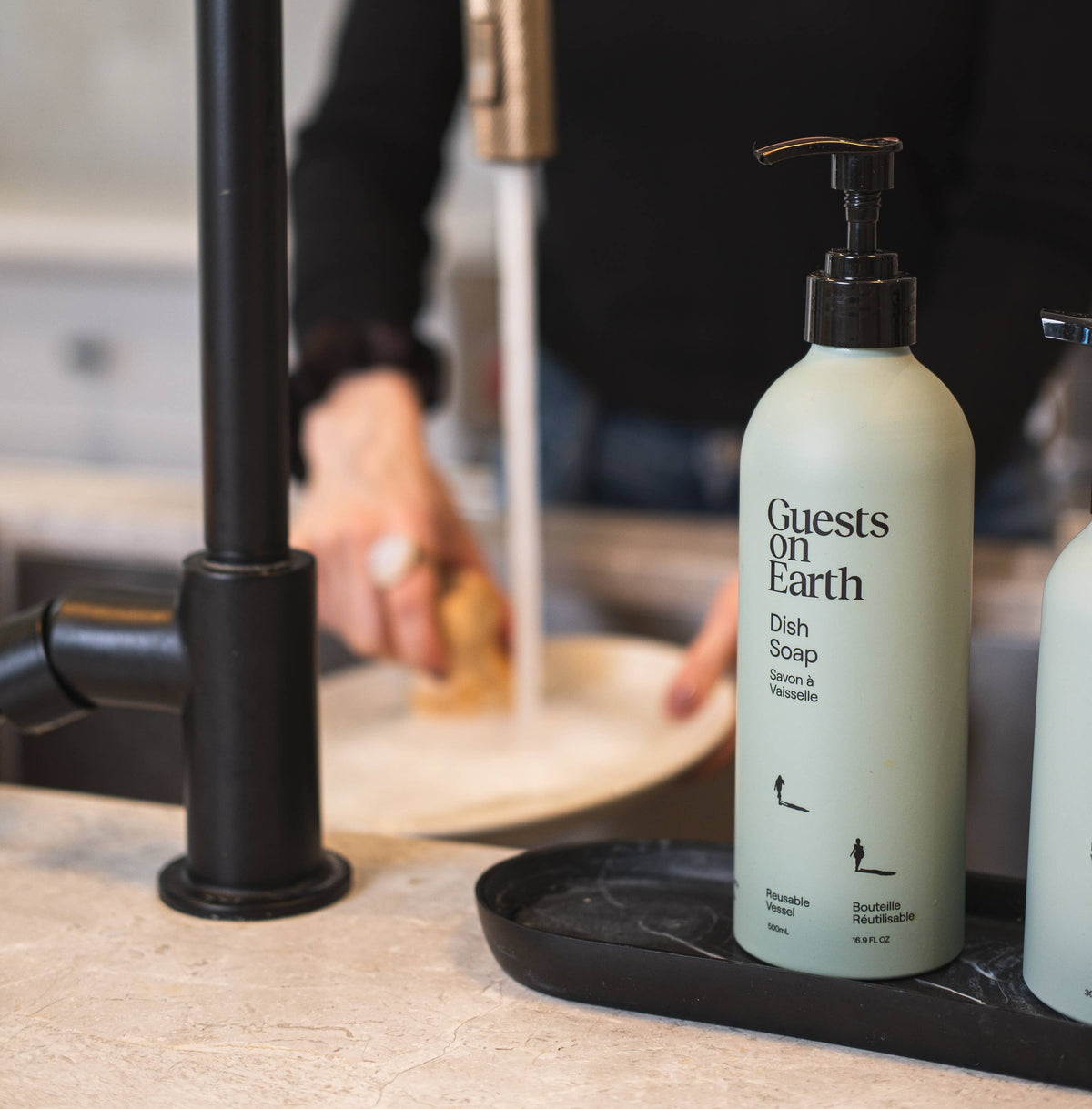 Guests on Earth dish soap pump bottle beside a kitchen sink while dishes are being washed.