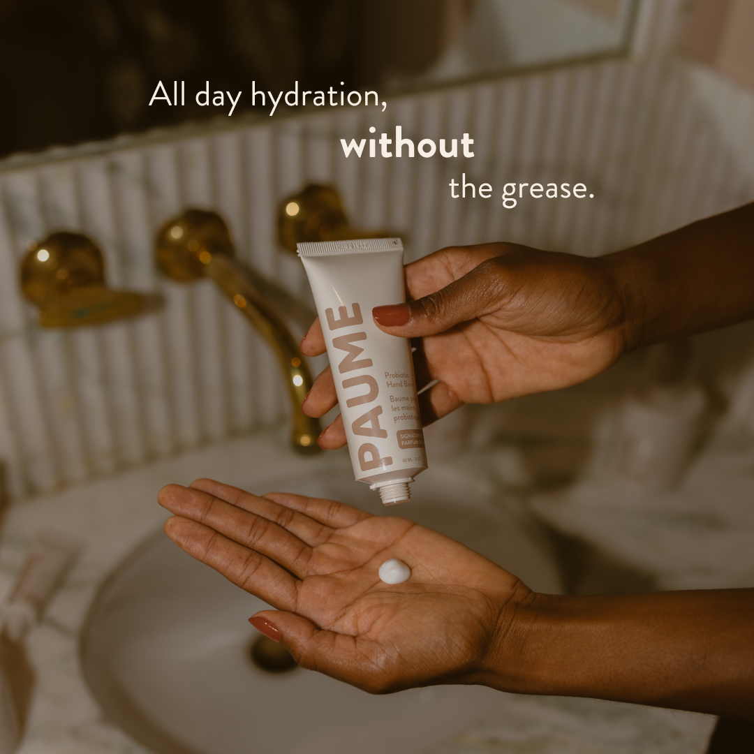Hand holding a tube of PAUME Hand Balm with text about hydration in a bathroom setting.