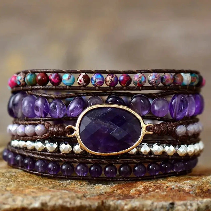 A stack of five multi-layered wrap bracelets displayed on a textured stone surface. The bracelets feature dark brown leather cords, purple amethyst beads, white pearl-like beads, a central oval gold bezel with a purple stone, and a top band with colorful marbled beads.