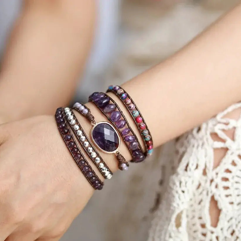 A close-up of a person's wrist wearing multiple stacked bracelets, including a central faceted purple gemstone bracelet, beaded brown and silver bracelets, and a floral-patterned bracelet, with a white lace garment in the background.
