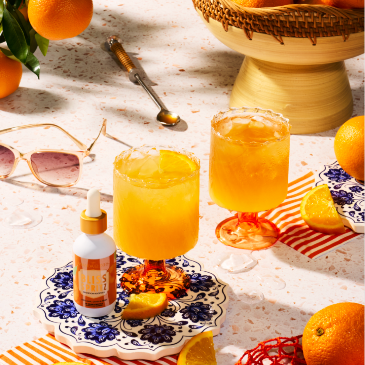 Two glasses of flavored water with a bottle of bliss elixir on a table with oranges and sunglasses.
