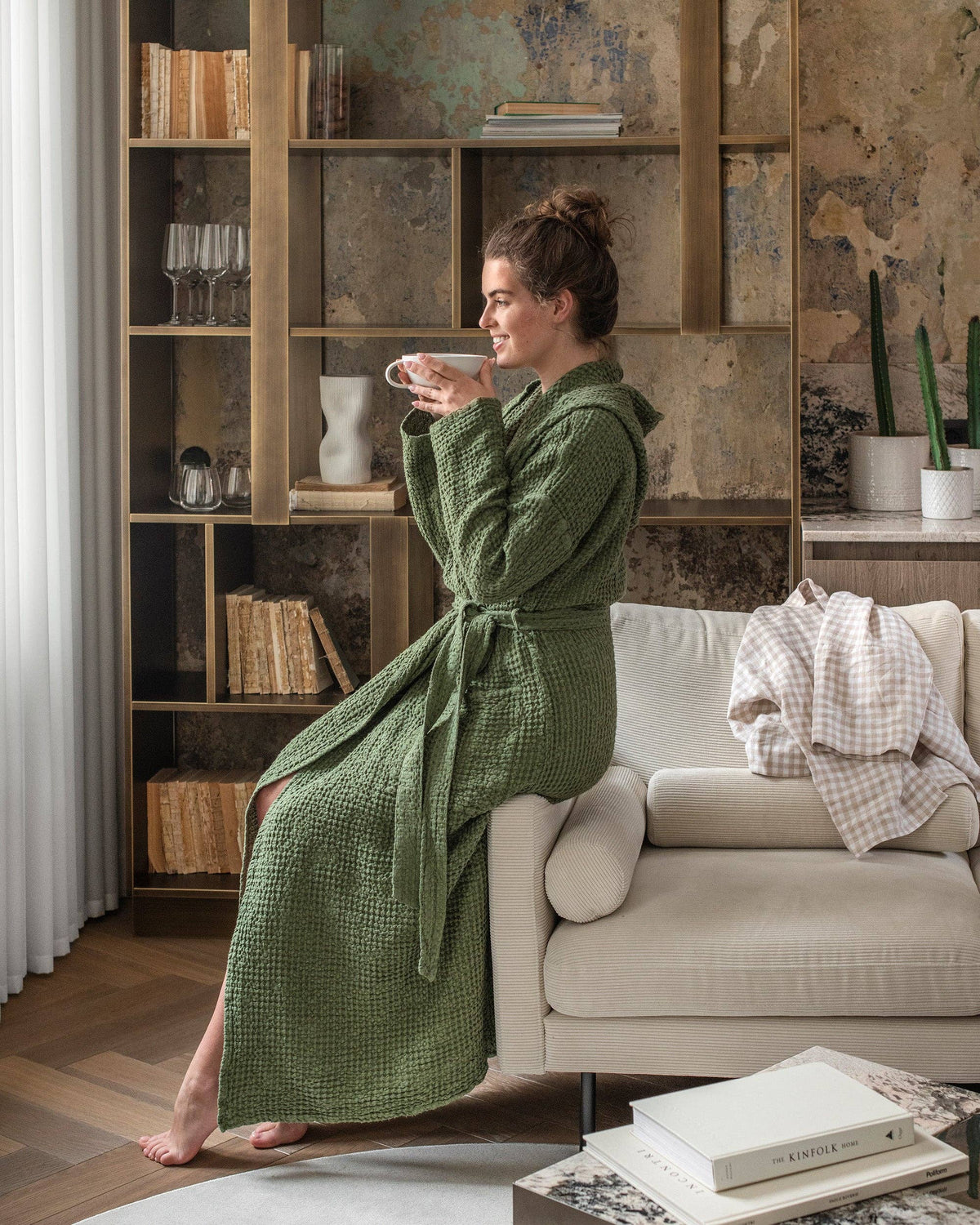 Woman in a green robe sitting on a couch in a cozy living room, holding a cup.