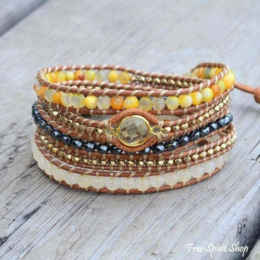 A stack of multi-strand wrap bracelets on a weathered gray wood background. The bracelets feature brown leather cords, yellow and orange beads, a black faceted bead strand, a cream-colored stone band, and a central gold-toned bezel setting with a clear crystal.