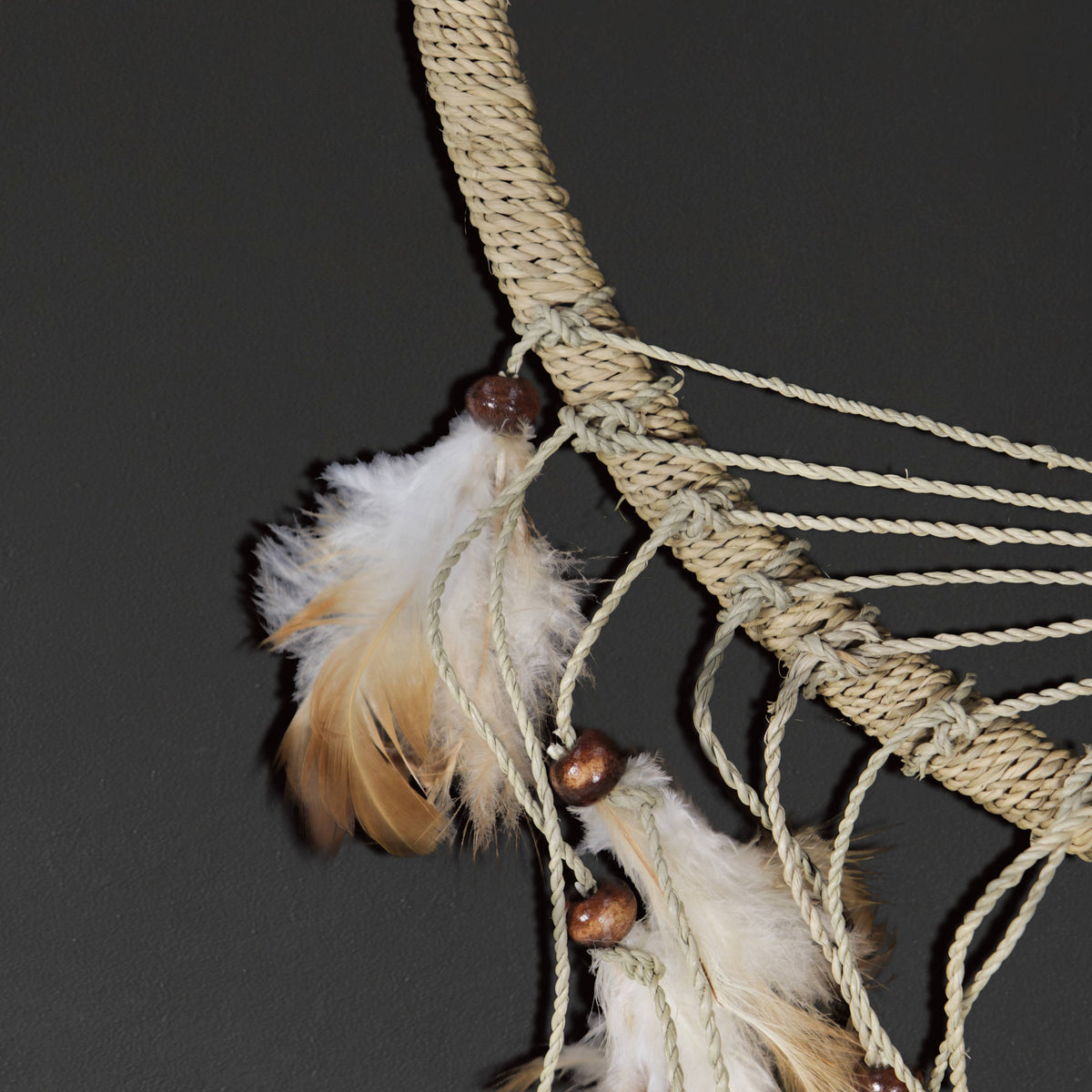 A close-up of a woven rope dreamcatcher with natural beige cord, white and tan feathers, and small brown wooden beads against a dark gray background.