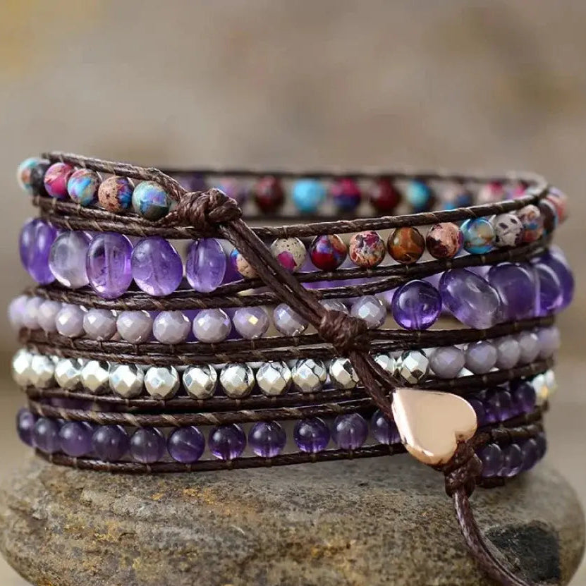 A stack of multi-layered wrap bracelets with purple beads, brown cord, iridescent beads, and a small rose gold heart charm, displayed on a stone with a blurred background.