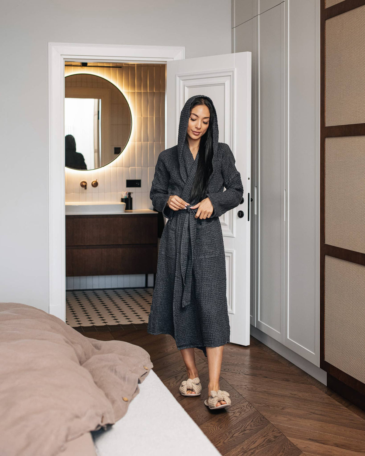 A woman wearing a dark gray textured robe and fluffy beige slippers stands in a doorway of a modern bedroom, looking down as if adjusting the robe.