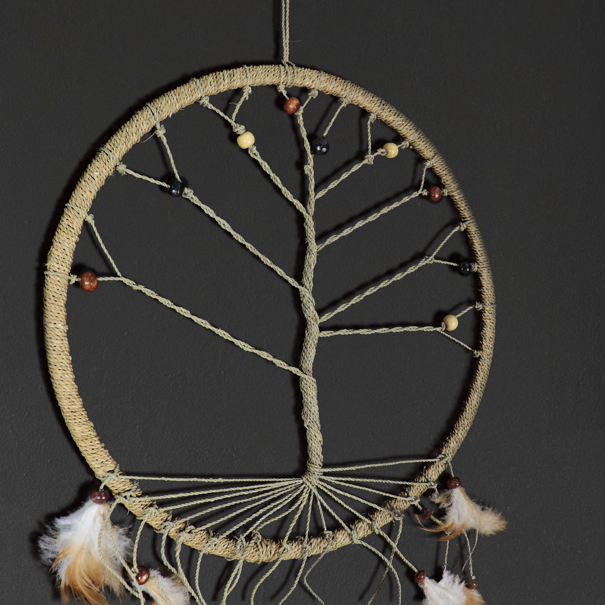 A circular dreamcatcher with a woven beige frame, featuring a tree design made of tan and brown cords, small wooden beads, and white feathers hanging from the bottom against a dark gray background.