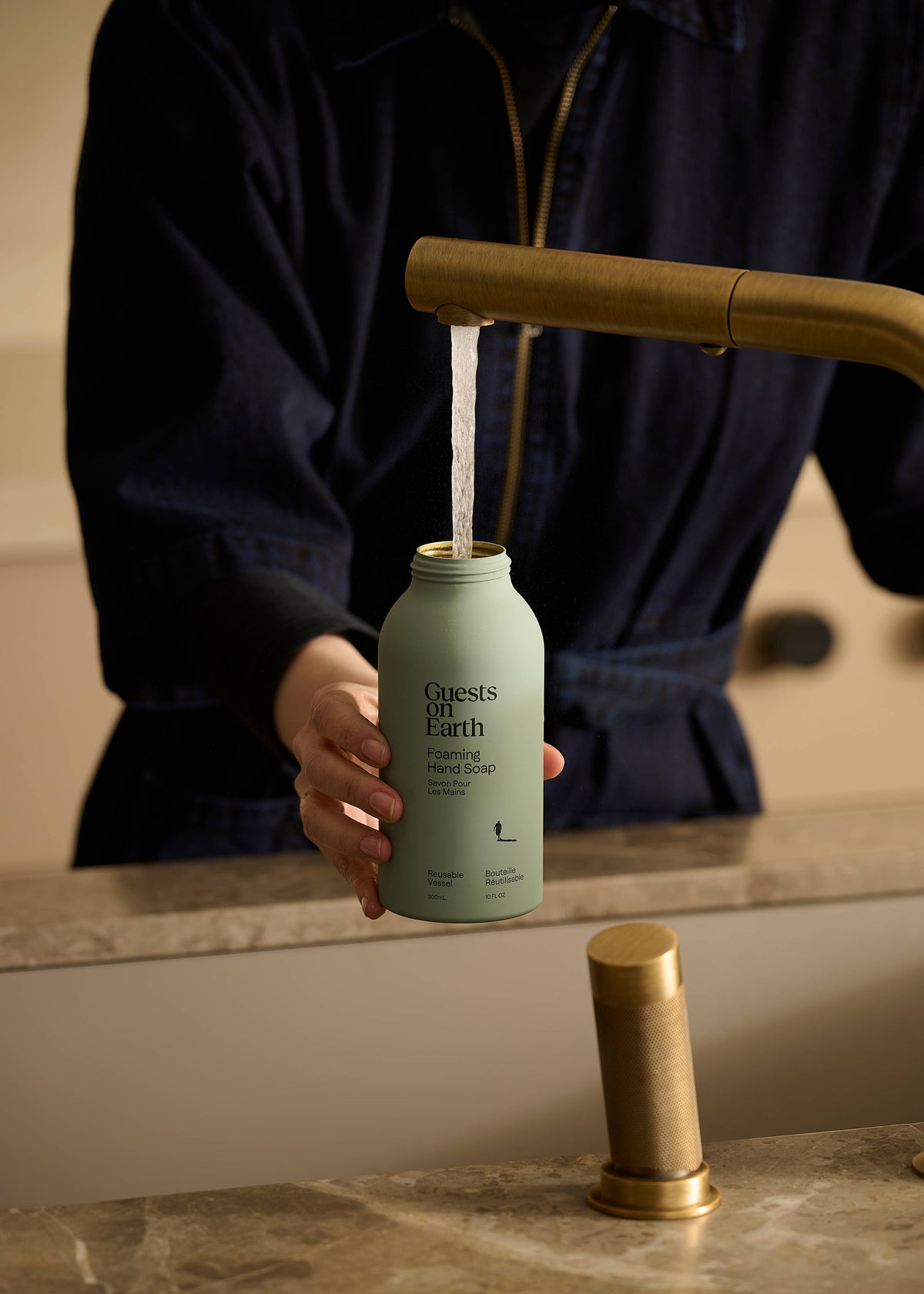 Person holding a Guests on Earth foaming hand soap bottle beneath a brass bathroom faucet.
