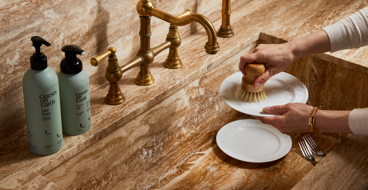 Person washing a plate at a brass kitchen faucet beside Guests on Earth pump bottles.