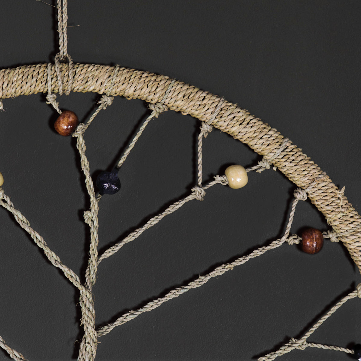 A close-up of a woven rope dreamcatcher with natural tan fibers, wooden beads in brown and cream, and a dark bead, set against a dark gray background.