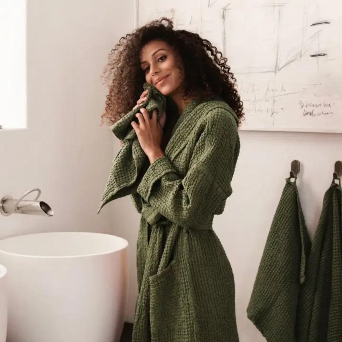 A woman with curly dark hair wearing an olive green waffle-knit bathrobe stands in a bright bathroom, holding a matching green towel. A white freestanding sink, wall hooks, and a sketch on the wall are visible.