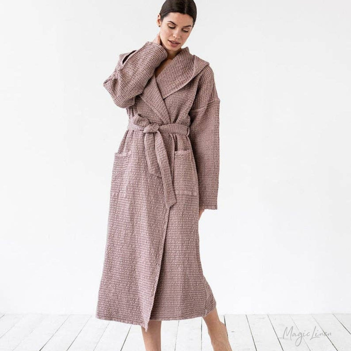 A woman wearing a long, textured waffle-knit bathrobe in a dusty mauve-pink color, standing against a clean white studio background.