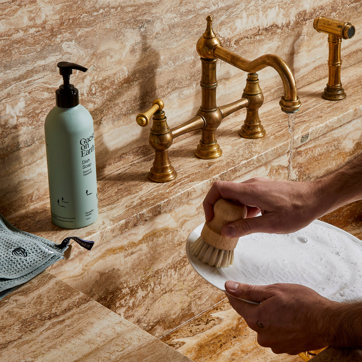 Person scrubbing a plate at a brass faucet beside a Guests on Earth dish soap bottle.