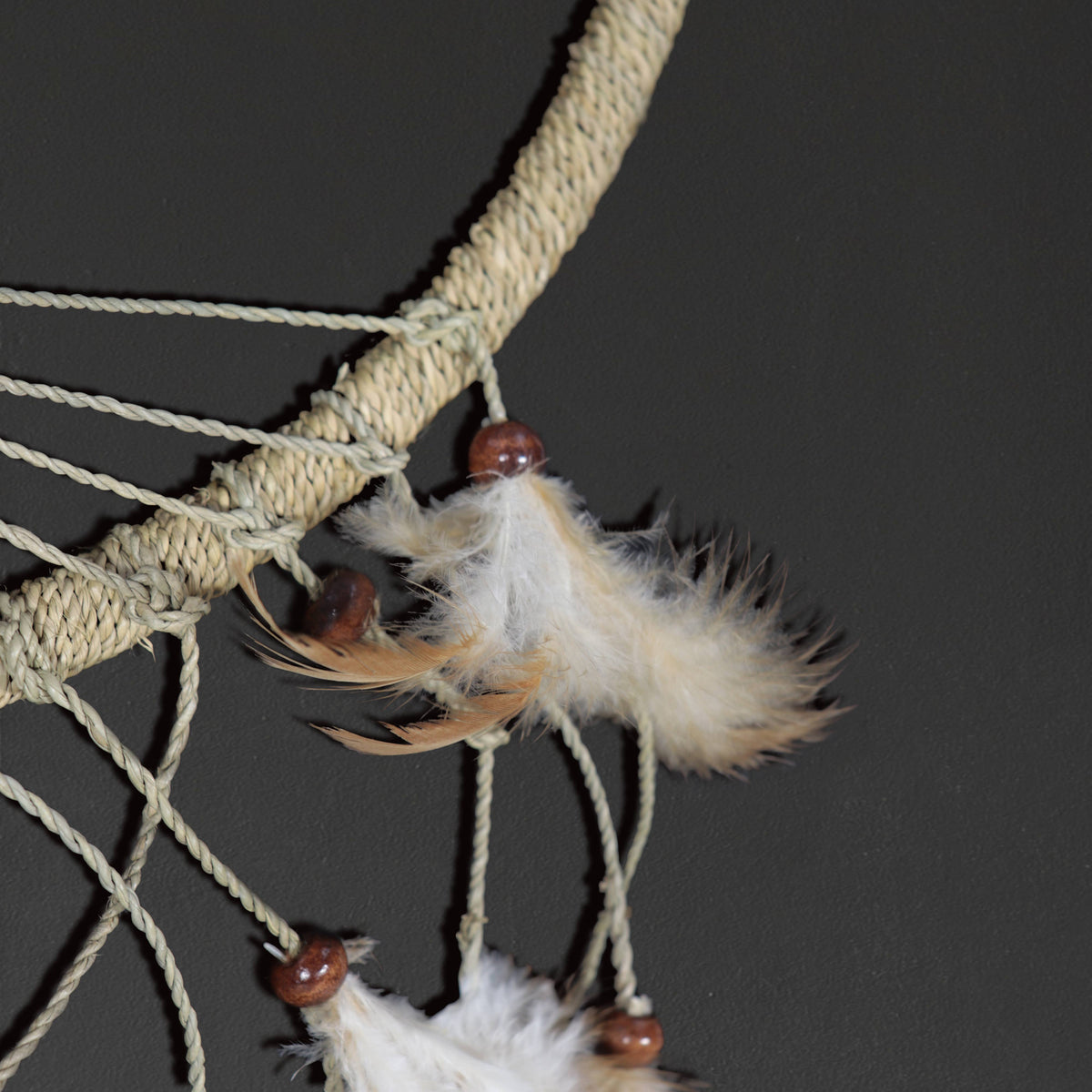 A close-up of a dreamcatcher with a thick beige wrapped branch, beige string webbing, white feathers, and small brown wooden beads against a dark gray background.