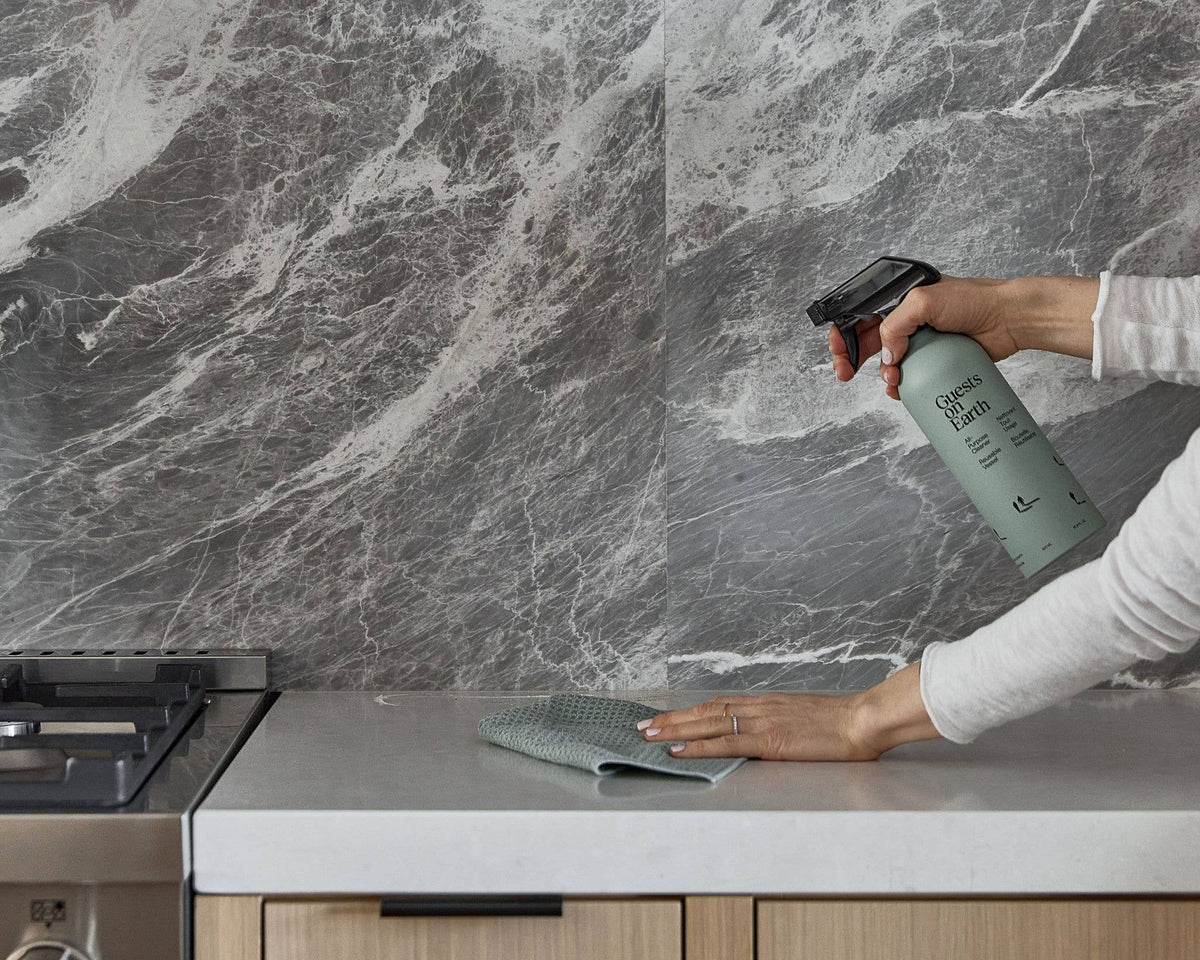 Person spraying Guests on Earth cleaner and wiping a kitchen counter beside a stovetop.