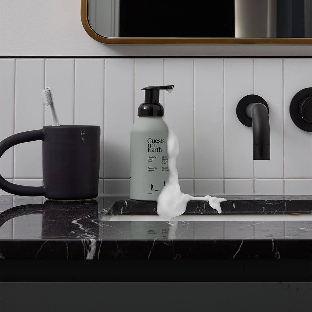 Guests on Earth foaming hand soap bottle beside a black bathroom faucet and white tiled wall.