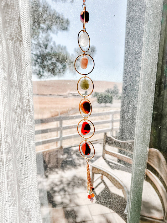 Seven Chakra Crystal Wall Hanging with colored stones in front of a window with a view of a snowy landscape.