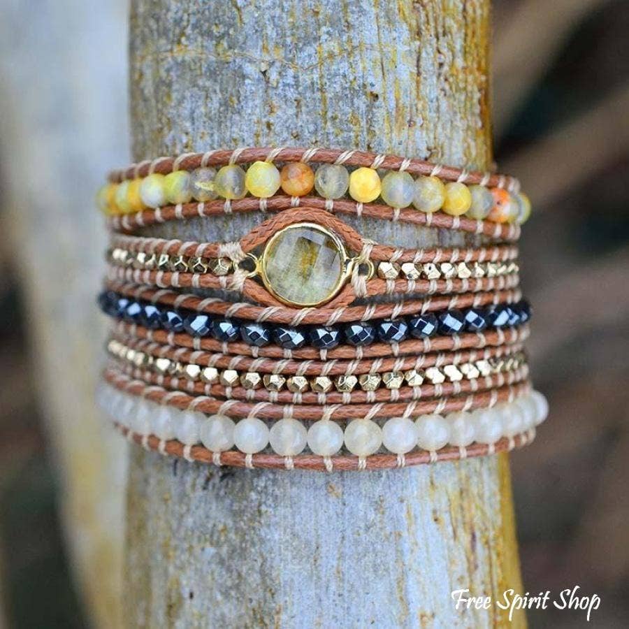 A stack of bohemian-style beaded bracelets wrapped around a weathered tree trunk. The bracelets feature natural wood beads in yellow, orange, and green, a central round stone charm, dark blue faceted beads, gold-toned metal spacers, and white beads.