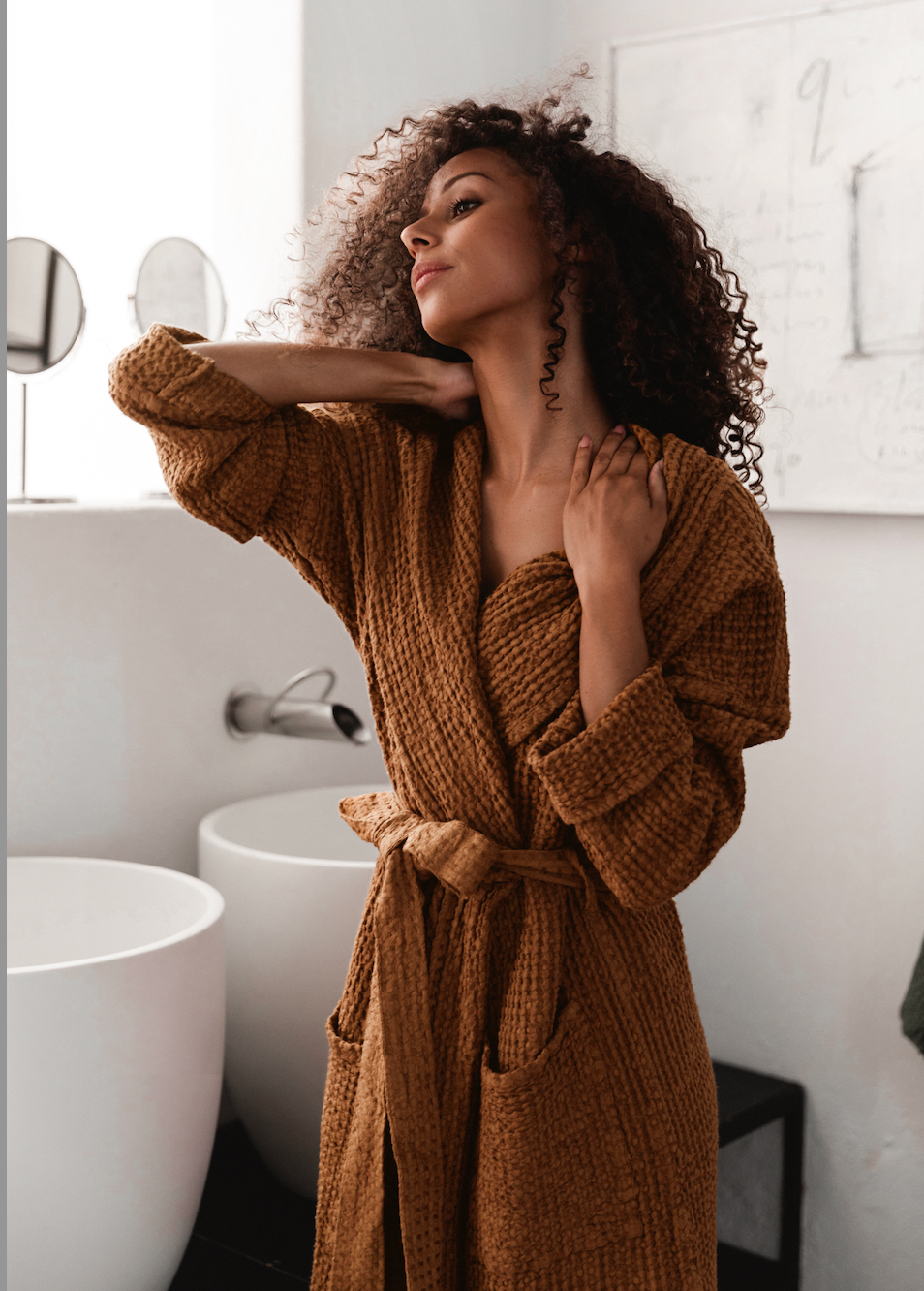 A woman with curly dark hair wearing a chunky waffle-knit brown robe, posing in a modern bathroom with white sinks and a whiteboard on the wall.