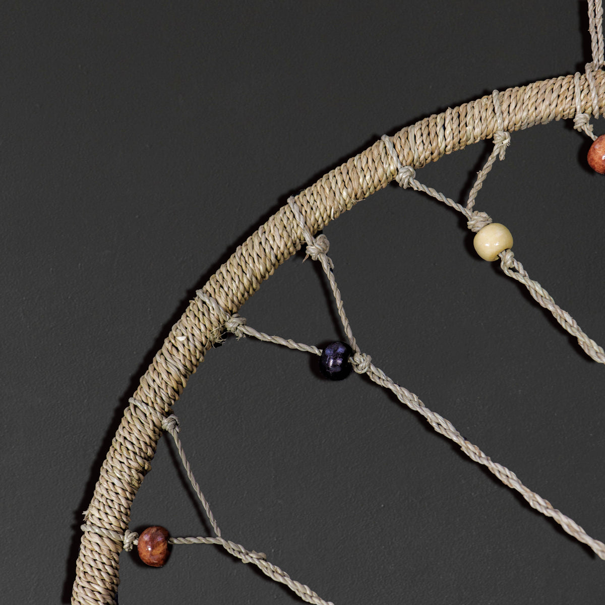 A close-up of a dreamcatcher featuring a thick, textured beige woven hoop and matching beige hanging strings adorned with small, polished beads in brown, purple, and cream, set against a dark gray background.