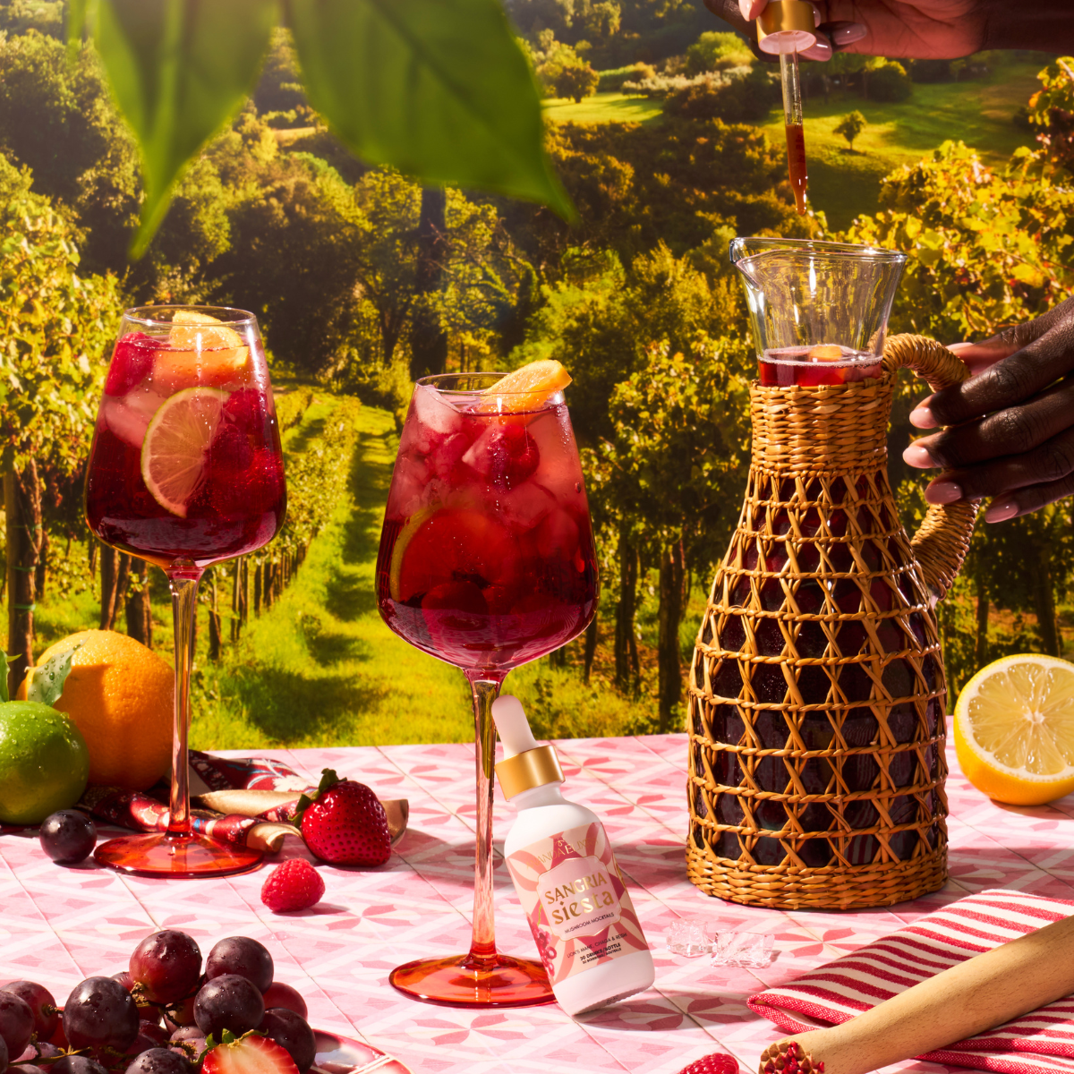 Two glasses of red sangria with fruit on a table outdoors, with a person pouring liquid into a pitcher.