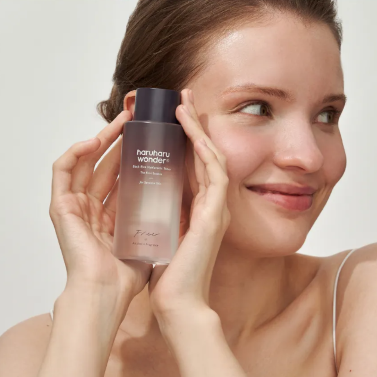 A woman holds a bottle of haruharu wonder - Black Rice Hyaluronic Toner Fragrance Free against her face, showing the product in use. The bottle is frosted with a light pink tint and has a dark cap.