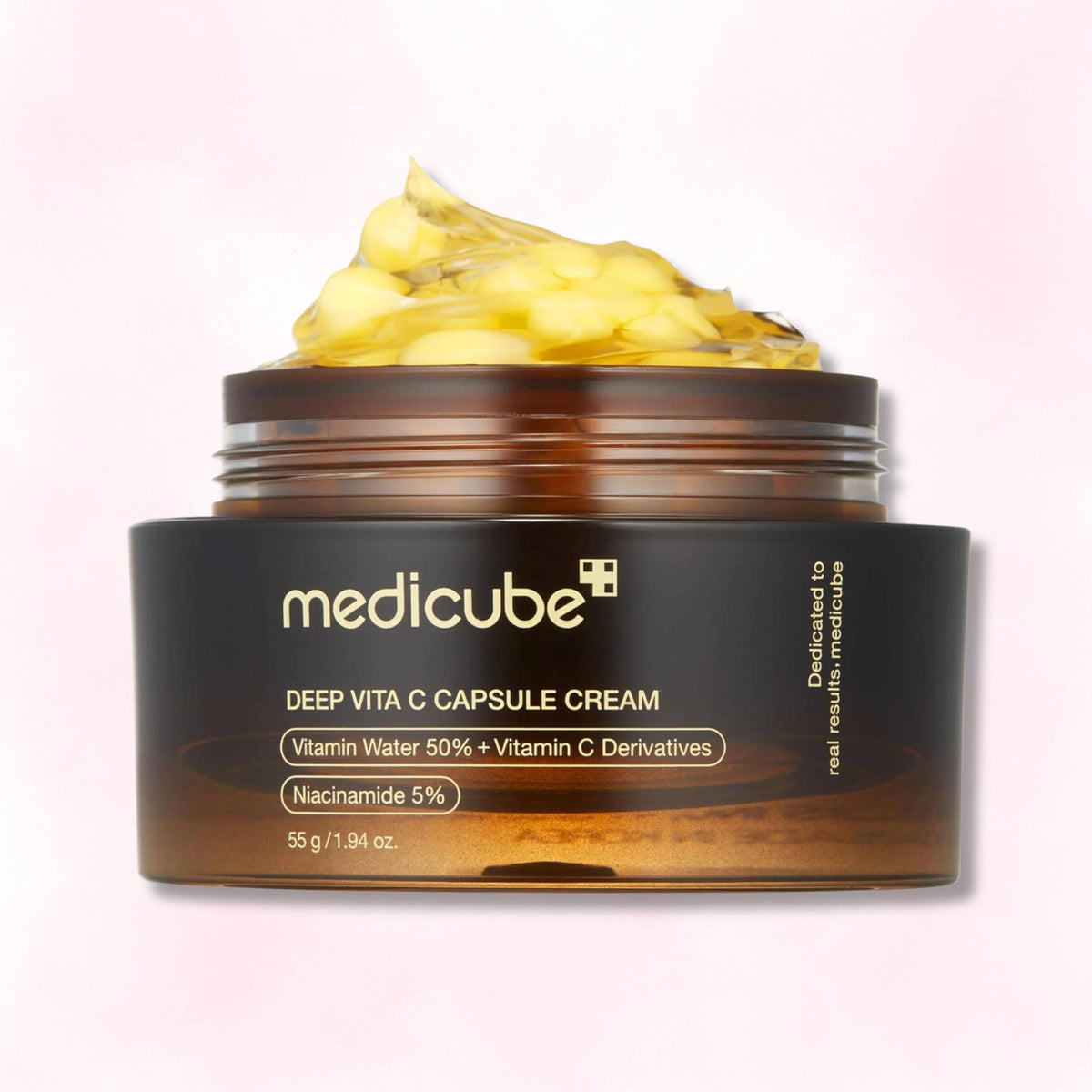 A jar of medicube Deep Vita C Capsule Cream with a dark brown label and amber jar, showing a dollop of pale yellow cream on top, set against a soft pink gradient background.