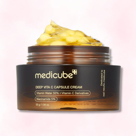 A jar of medicube Deep Vita C Capsule Cream with a dark brown label and amber jar, showing a dollop of pale yellow cream on top, set against a soft pink gradient background.